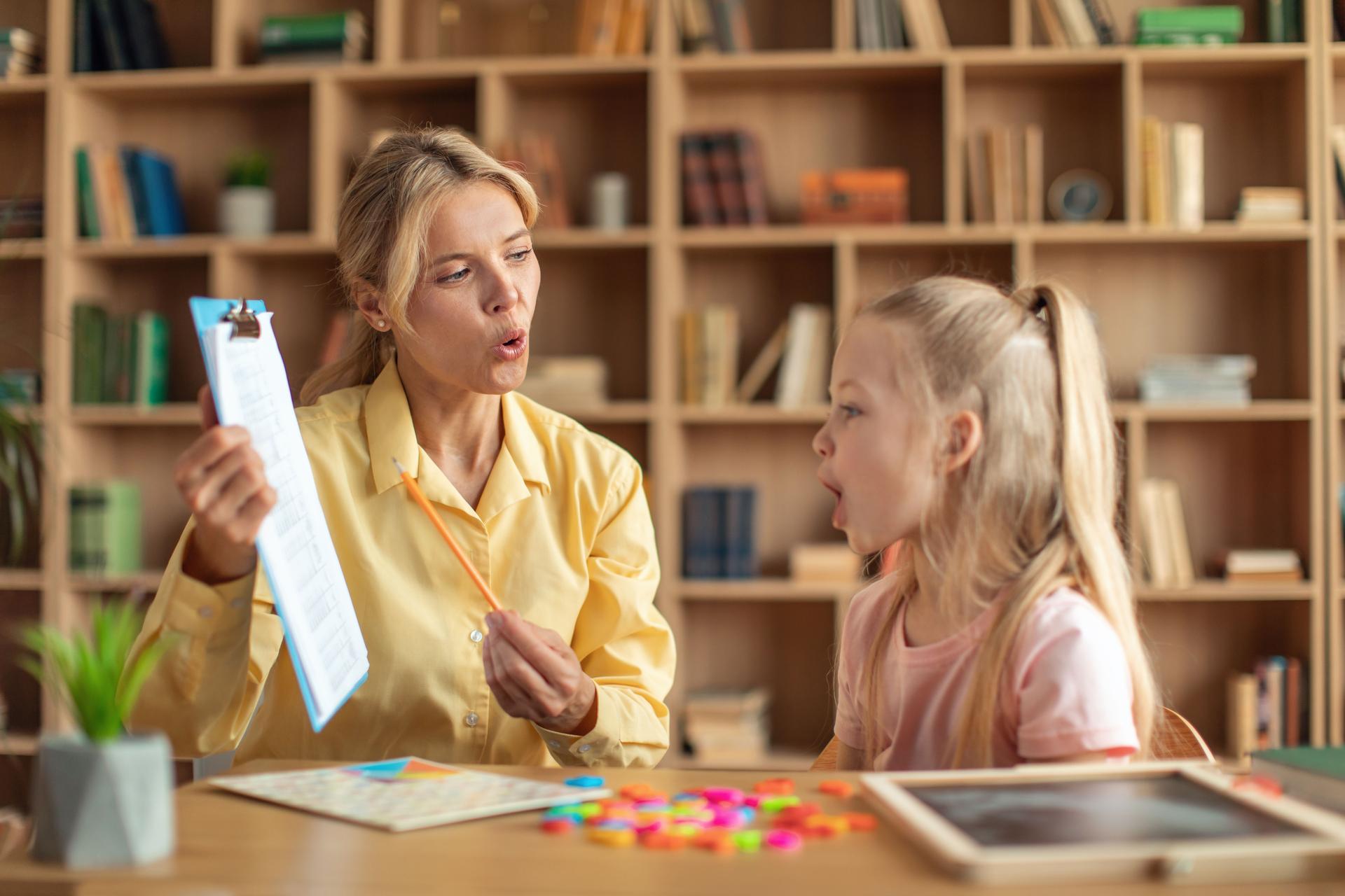 Cheerful female speech therapist curing girl's problems and impediments, cute child learning and pronouncing letters